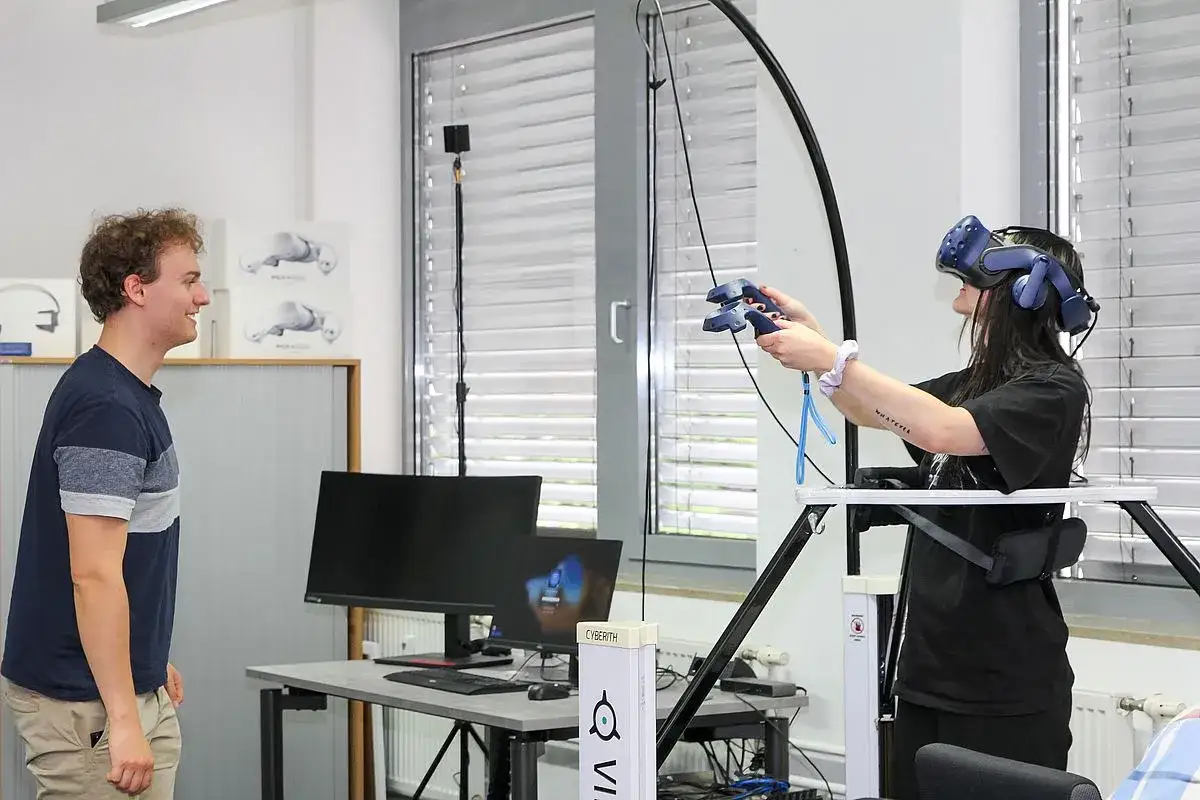 Ein Mann beobachtet eine Frau, die eine VR-Brille trägt und Controller hält. Hier wird gelebt, was ist Medieninformatik: Interaktion mit virtuellen Welten.