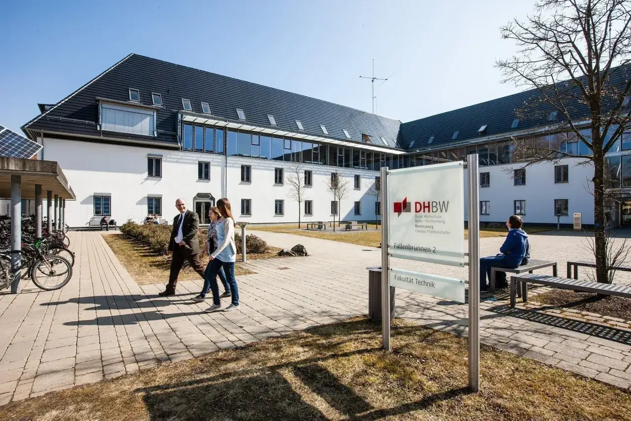 Sonniger Tag am Campus der Hochschule Friedrichshafen. Menschen gehen &uuml;ber den gepflasterten Weg vor dem modernen Geb&auml;ude.