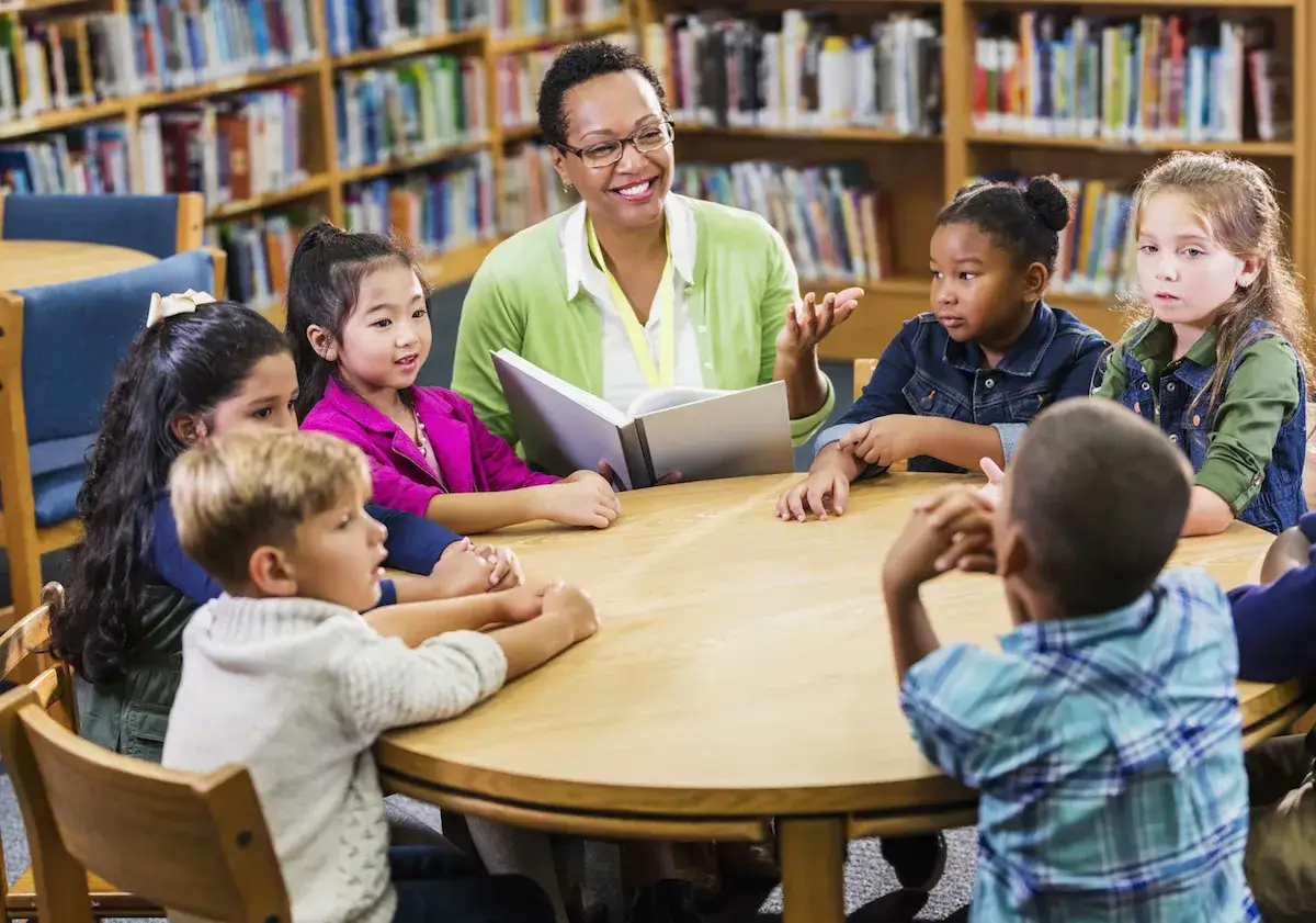 Lehrerin liest Kindern im Bücherregal vor. Die Kinder lauschen gespannt, was das Lesen für sie bedeutet.