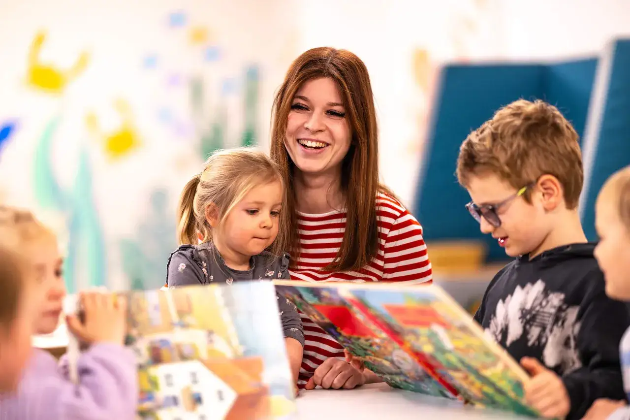 Eine junge Frau mit roten Haaren liest Kindern vor. Sie lächelt fröhlich. Die Kinder sind aufmerksam. Vielleicht denkt sie über ihre PIA Ausbildung und das Gehalt netto nach.